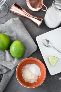 a small bowl that has sugar and shredded coconut next to 2 limes and a plate