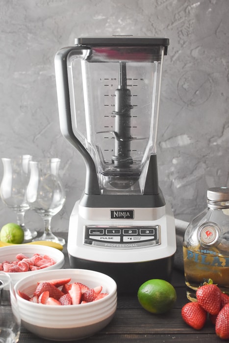 An empty blender next to all of the ingredients for a frozen strawberry daiquir
