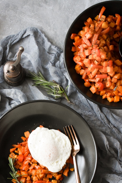 Two black bowls, one with roasted butternut squash and peppers, the other with the same mixture topped with an egg.