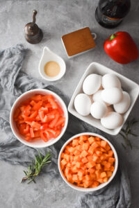 An ingredient shot that includes eggs, red bell peppers, chopped butternut squash, salt, pepper, garlic and fresh rosemary.