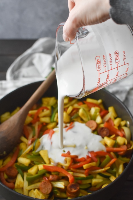 Adding coconut milk to a pan of cooked sausages and vegetables.