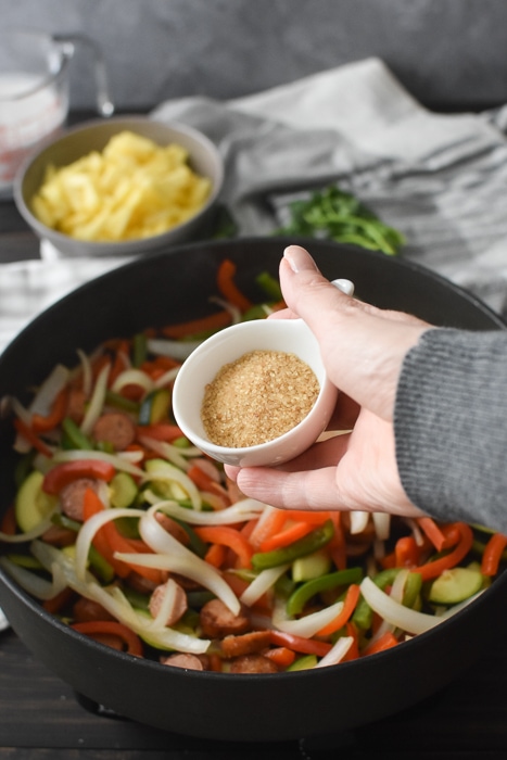 Adding sugar to a pan of cooked sausage and vegetables.