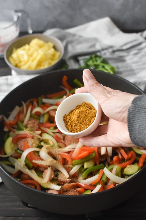 Adding curry powder to a pan of cooked sausages and vegetables.