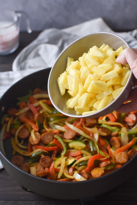 Adding fresh pineapple to a pan of cooked sausages and vegetables.