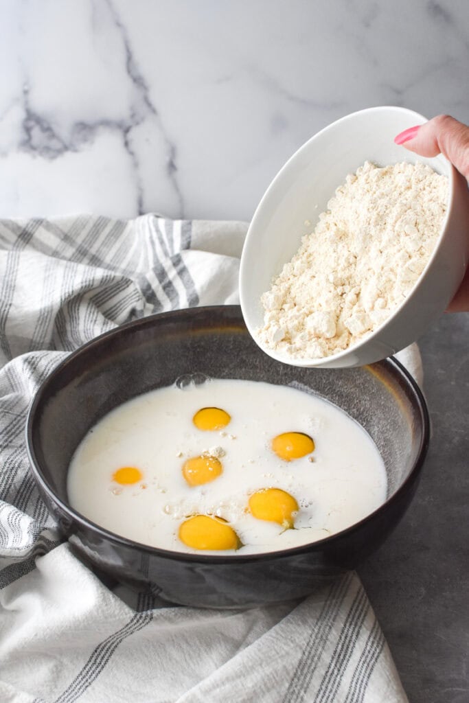 Adding flour to the ingredients for Whole Wheat German Pancake in a large brown bowl.